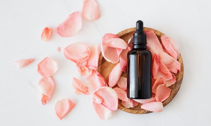 serum in a dark bottle in a wooden bowl full of pink rose petals on a white background