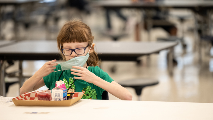 food for school little girl with glasses and a mask dressed in a green shirt sitting alone in front of a lunch tray at school