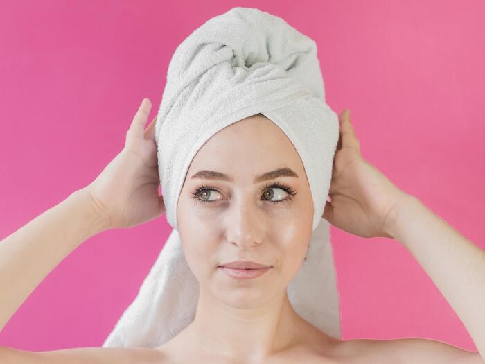 woman with a white towel on her hair face close up on a bright pink background holding her head with hands