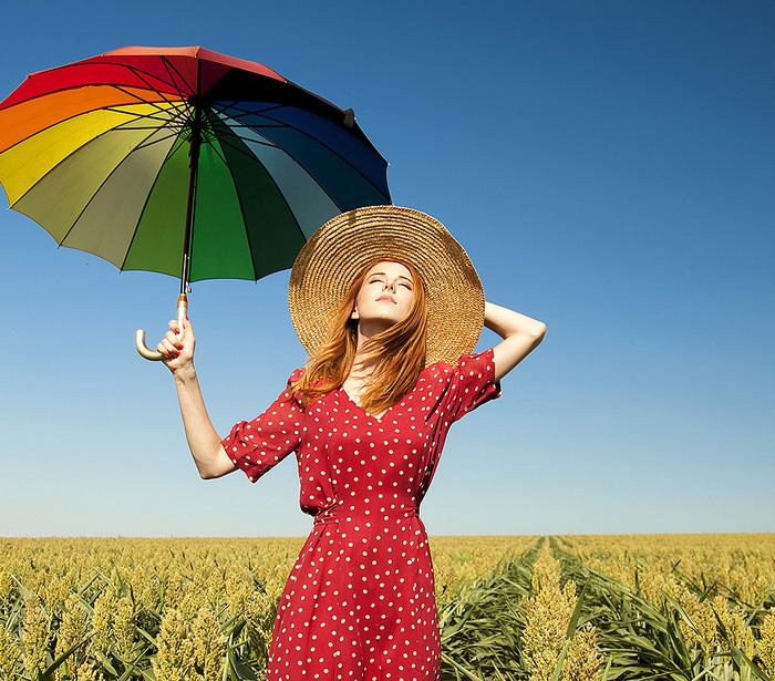 red-haired woman in a dotted red dress in a field with a sun hat and a large colorful umbrella looking at the soon with her eyes closed