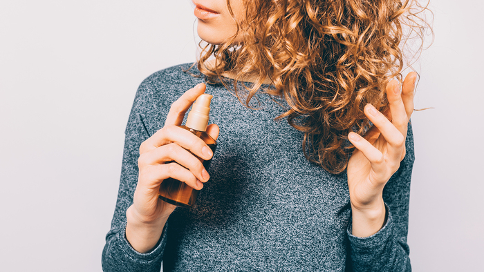 woman with curly hair dressed in a grey sweather applying protective hair spray to her curls