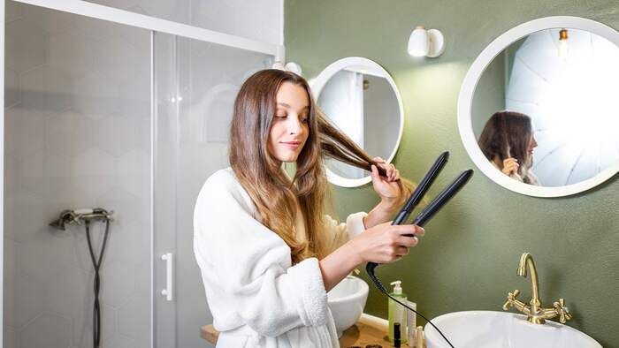 woman in a white bathrobe in front of two mirror in the bathroom styling her hair with a hairpress