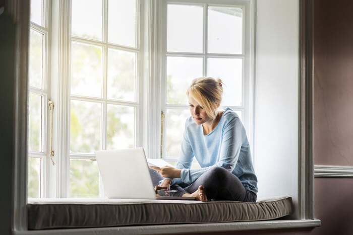 indoor sunscreen blond woman with her hair up in a light blue blouse sitting in a wide window sill in front of a large window and a white laptop