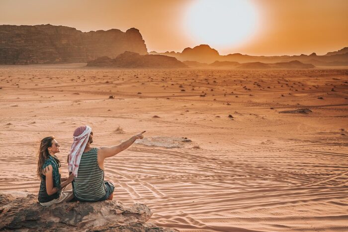 jordan eco tours woman and man sitting on a rock in the desert man with a scarf on his head pointing towards the horizon