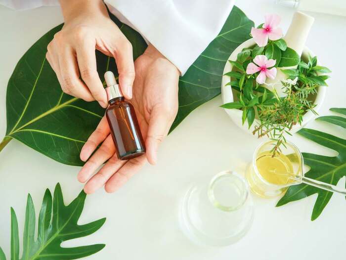 organic serum hand holding a dark serum bottle surrounded by green plants and flowers