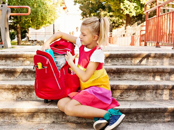 packing for school little blond girl in colorful dress sitting on the stairs packing a large red backpack for school