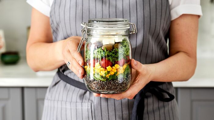 woman dressed in grey apron in a kitchen holding a large jar full of vegetables and seeds