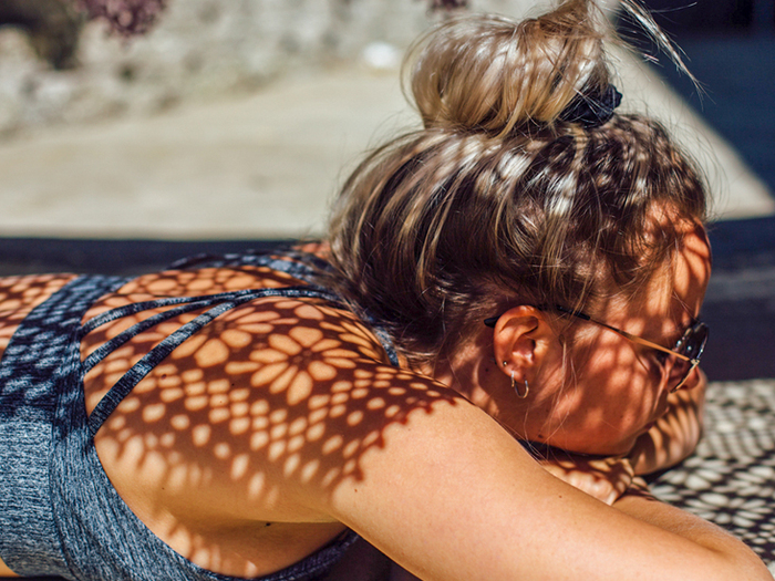 blond woman with her hair up and sunglasses in a blue top lying in the sun with some shadow patterns on her back