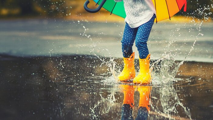 child with yellow boots blue pants and a white sweater holding large colorful umbrella and playing in the rain on the street