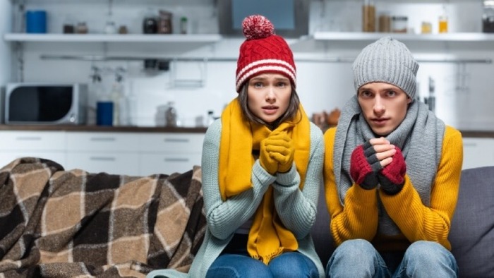 couple sitting on a couch in their kitchen with scarves and hats on being cold at home