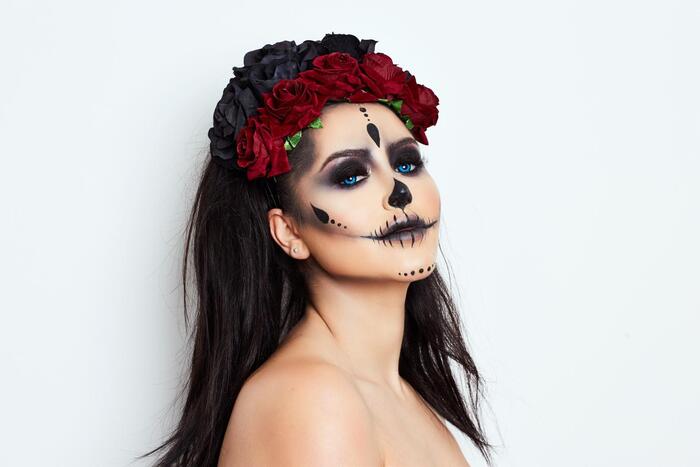 woman with black hair posing on a white background with halloween make up on and a crown of red and black roses