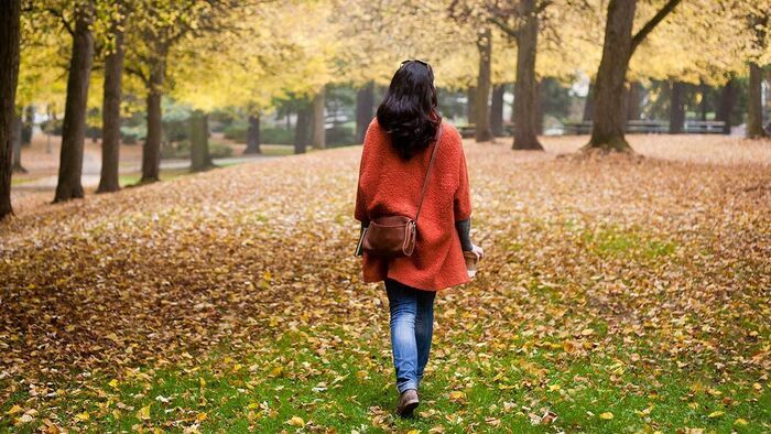 woman dressed in jeans and an orange coat walking in the park with a brown cross body bag and her hair loose