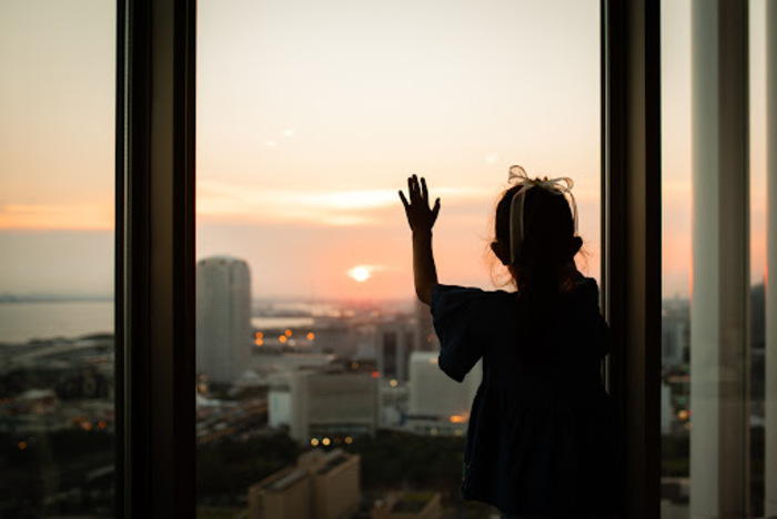 little Girl with a ribbon in her hair watching the sunset from the window of the apartment