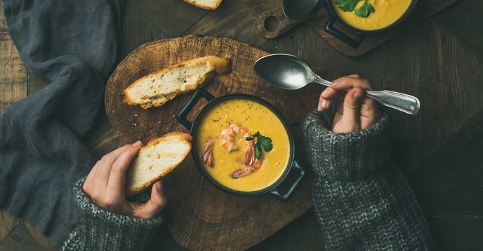 hearty meal woman in a grey sweater eating a thick yellow soup from a black bowl with toast