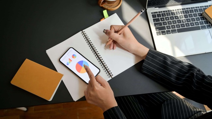 phone and writing woman in a suit in front of a dark desk with a laptop notebook and phone checking some graphs