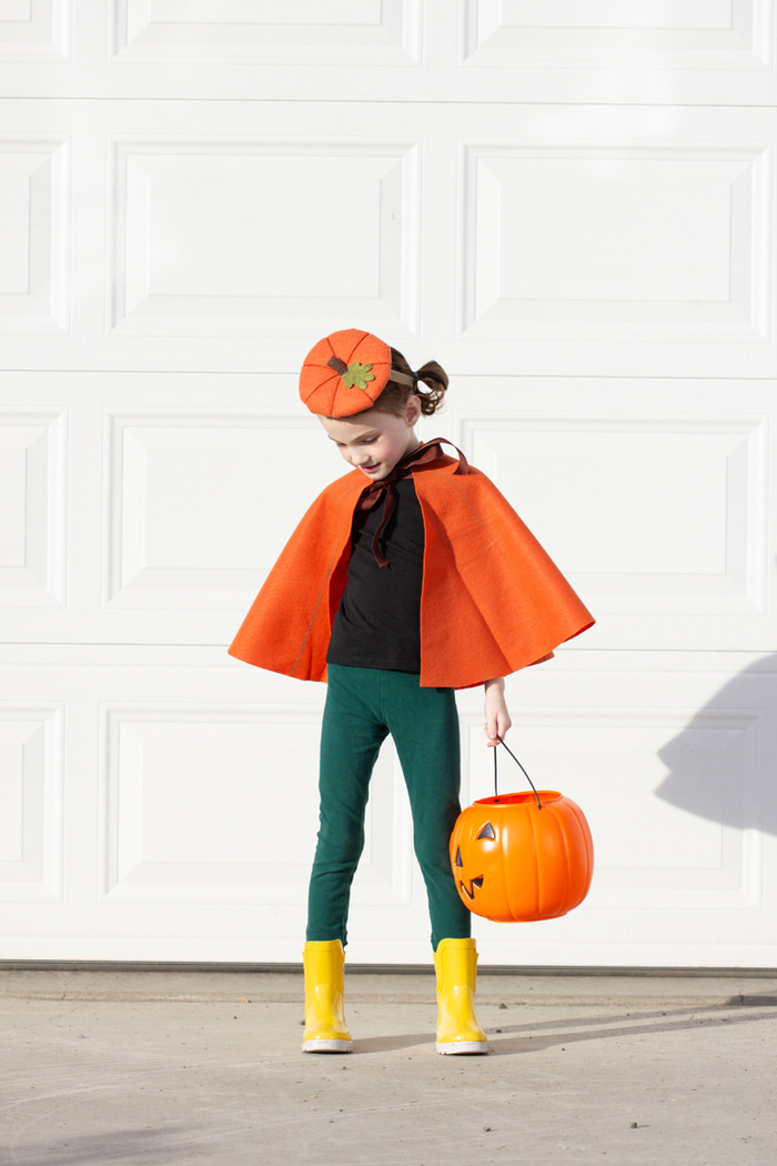 pumpkin cape halloween costume little girl in front of a white wall in a fall inspired costume with a pumpkin lantern