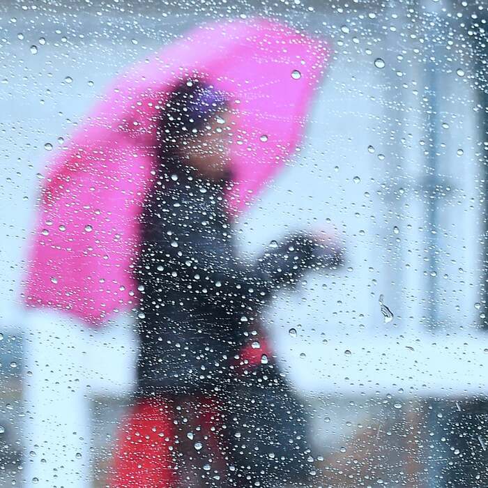 window with rain drops on it and a woman dressed in a dark jacket and with a large pink umbrella outside