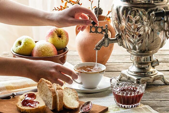 woman pouring russian tea from a large pot in a white tea cup with slices of bread jam and apples in the background