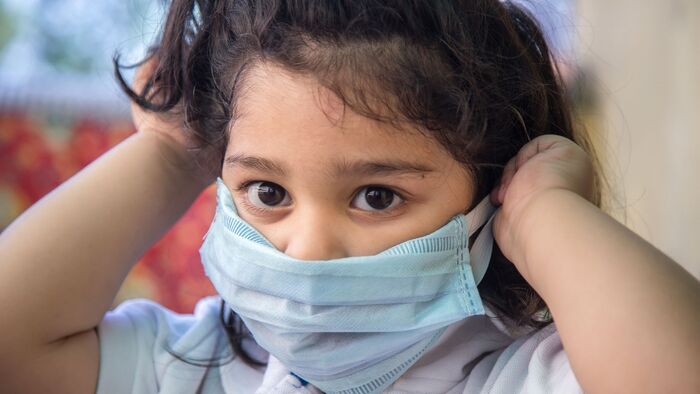 child with dark eyes and dark hair holding up a blue mask in front of their mouth