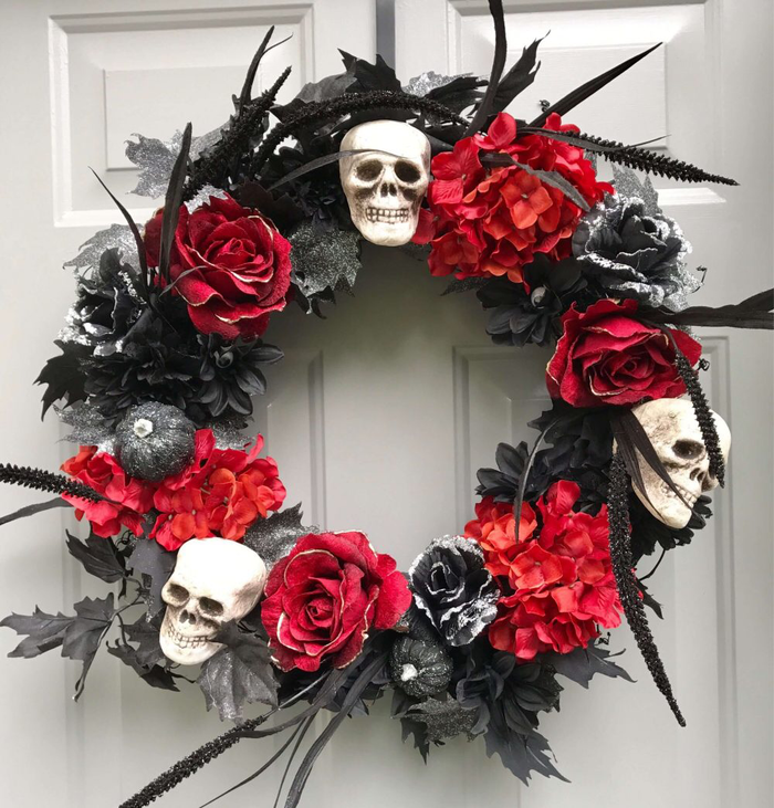 spooky faux flower wreath in red and black with white skulls black feathers and red roses hanging on a white wall