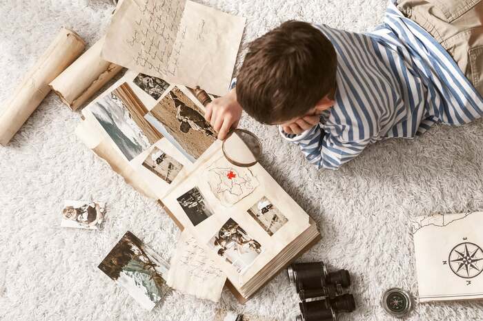 little boy lying on the floor on a fluffy carpet with a large photo album open in front of him with a magnifying glass looking at pictures