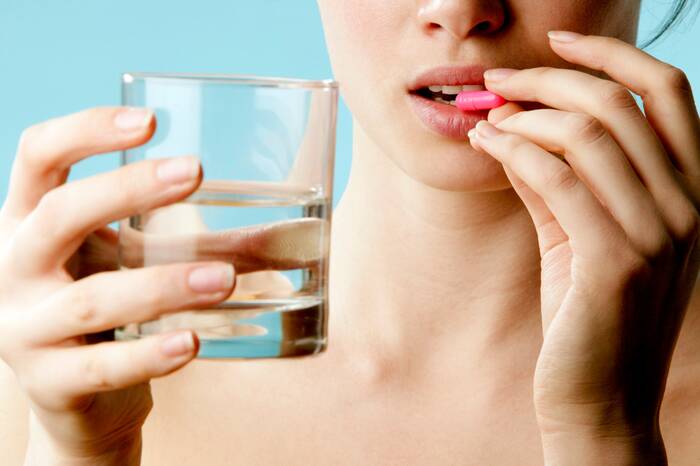 woman holding a glass of water with a pink pill in her hand close to her lips