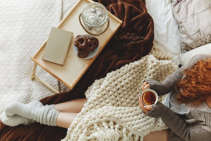 red-haired woman with a warm drink in her hands in bed with wool socks on and two thick blankets in front of a serving tray