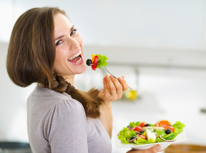 woman dressed in a grey top holding a bowl of salad eating and looking back at the camera
