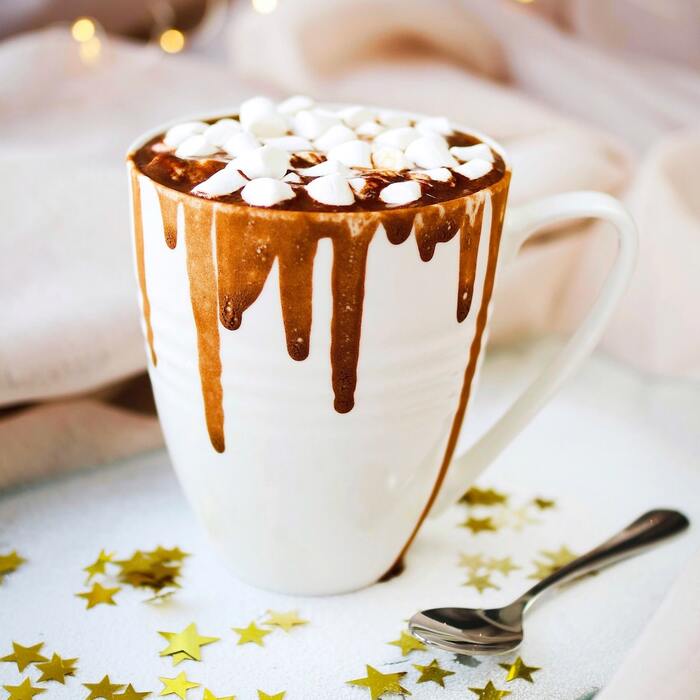 large white mug with hot cocoa dripping out filled with small marshmellows with a spoon and stars 