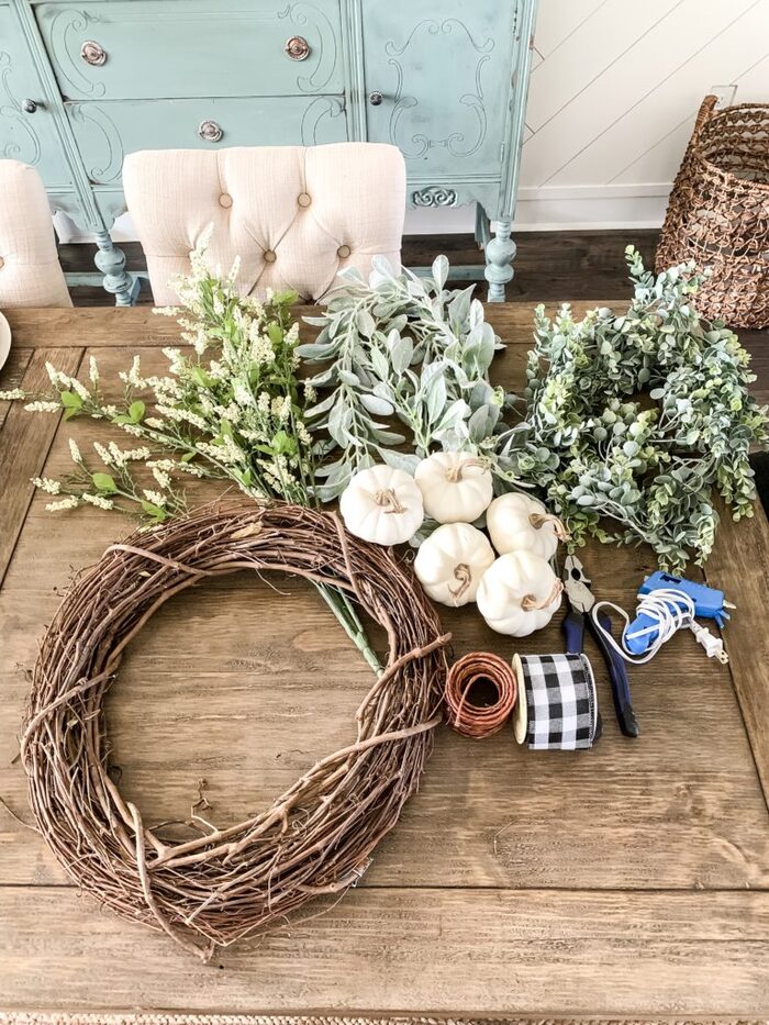 DIY materials lying on a large wooden table in a living room branches white pumpkins and a wreath base of branches