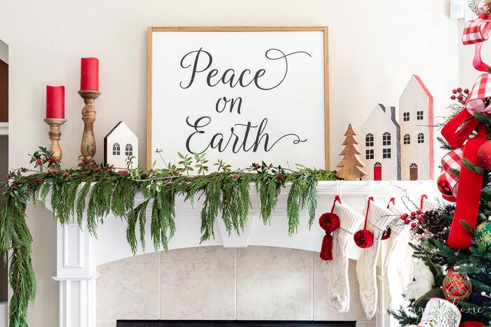Christmas home decor on a mantel with green foliage and a sign peace of earth next to wooden candle holders and little houses