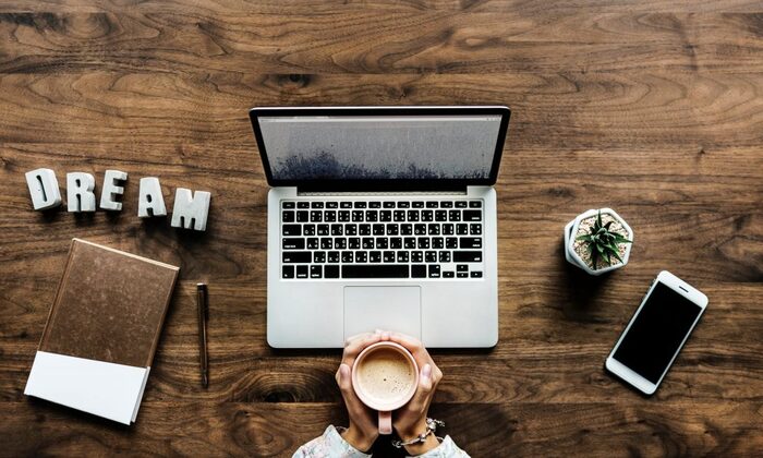 woman holding a cup of coffee with both her hands in front of a computer on a wooden table with a phone and notebook next to her