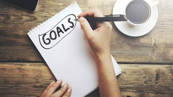 woman writing down her goals on a white notebook with a cup of coffee next to it on a wooden table