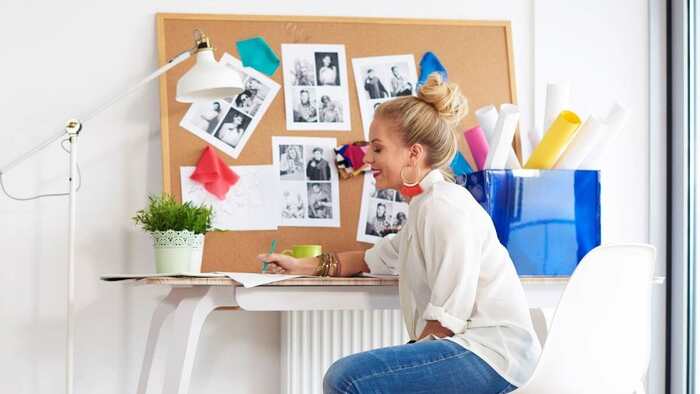 blond woman in jeans and white shirt sitting on her desk designing a vision board with photos in front of her