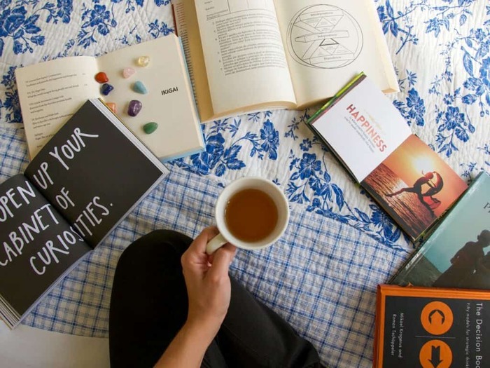 woman holding a cup of tea on a bed with books scattered around her