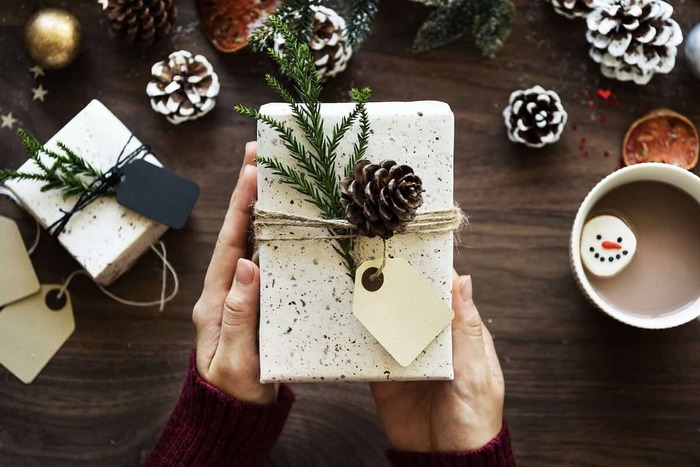 womans hands holding a pretty gift wrapped in white paper with pinecones and other decorations scattered in the background