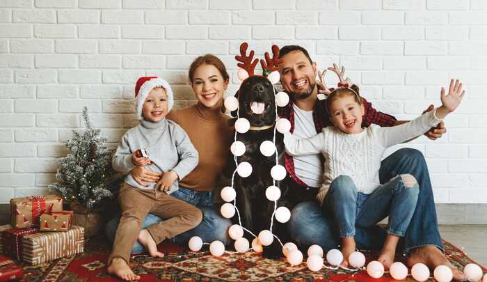 holiday family family of four sitting on the floor with a white brick wall background with christmas hats and a black dog smiling 