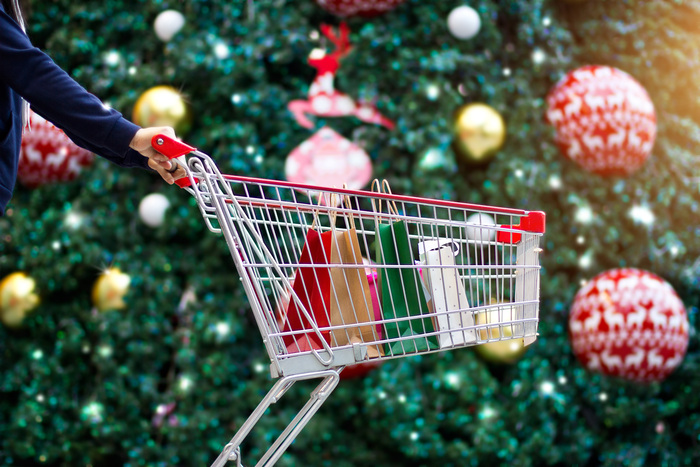 woman pushing a shopping cart with christmas gifts in it in front of a giant christmas tree