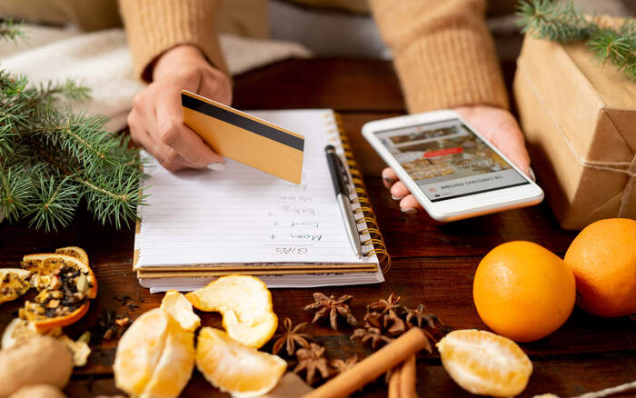 Woman holidng her phone and credit card with a notebook surrounded by christmas decorations planning and budgeting