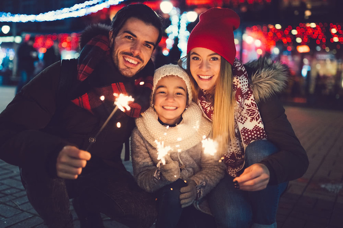 a family of three outside in a city dressed warm holding sparklers and smiling at the camera