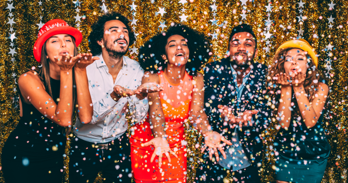 friends dressed in festive clothes in front of a sparkling wall laughing and spreading glitter