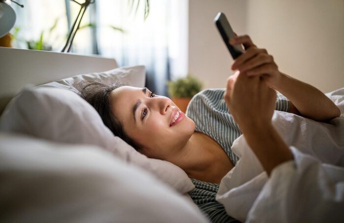 woman lying in bed smiling at her phone and looking at the screen