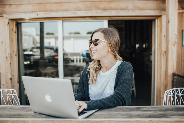 woman with sunglasses sitting outdoors smiling and working on her laptop