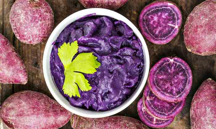 purple foods purple potato puree in a white bowl with a green veggie decor and slices of purple potatoes around