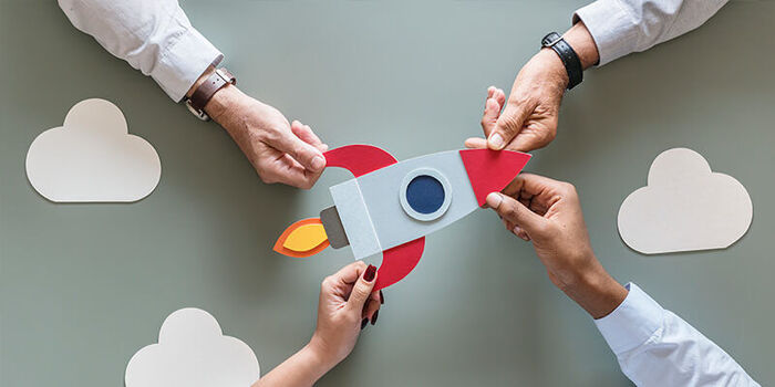 four hands holding a rocket on a gray background with white cut out clouds around