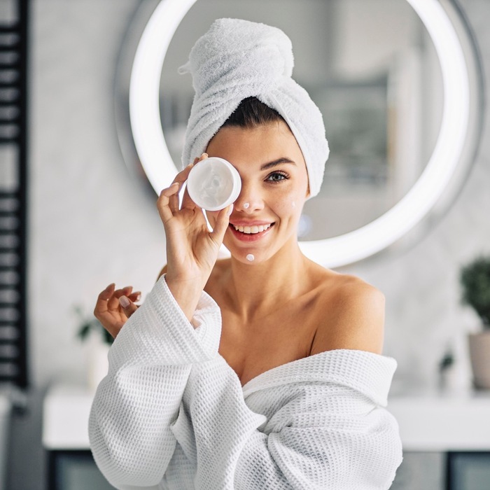 woman in a bathroom with a large window in the background with a white towel on her hair applying cream on her face