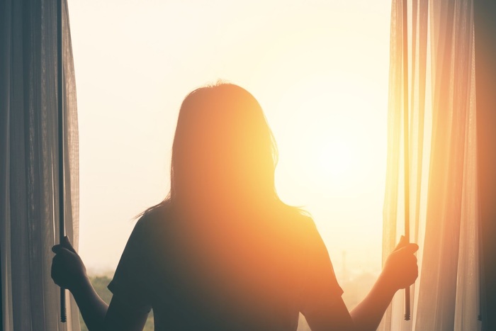 woman with her back to the camera opening the curtains of her window letting the sunlight in and looking outside