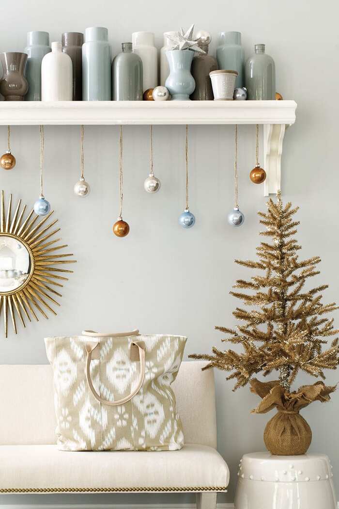 colorful christmas baubles handing from a shelve with vases of different size on it in a dove gray colored interior above a small couch with a golden christmas tree 