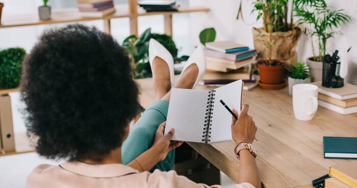 woman sitting with her back to the camera and her legs up on a desk with a notebook in her hands writing down some goals