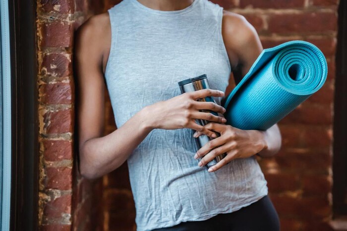 woman person leaning on a brick wall in sportswear holding a water bottle and a yoga mat